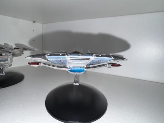 A model of a futuristic spaceship displayed on a black stand, with a sleek design and blue and red accents. The background features a white shelf, suggesting it's part of a collection.