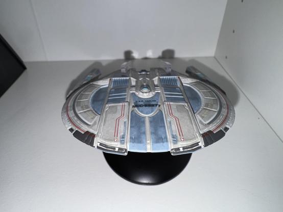 A model of a futuristic spaceship displayed on a black stand, featuring a sleek design with blue and silver colors and intricate details. It is placed against a white background.