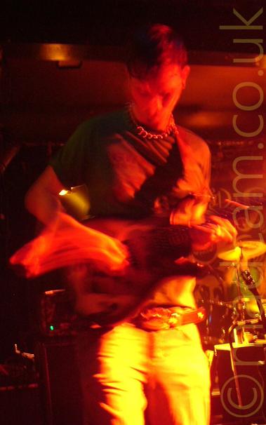 A man with short dark hair, wearing a green t-shirt and light-coloured trousers, playing an electric guitar on a stage.
A long exposure and red stage lights conspire to flood the image with red and yellow light, showing off the movement of his arms and guitar.
A drumkit can be seen behind him on the right, with amplifiers in the background on the left., with dark walls and ceiling with a red cast filling the rest of the frame.