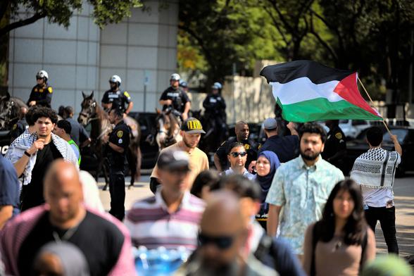 A shot of a crowd of protestors crossing the street after being forced to move by the police as their location by the building was deemed hazardous. The subject is on the right: a man holding a Palestine flag and wearing a kuffiyeh. His back is turned to the camera as he looks upon the horse-mounted police that were called in.