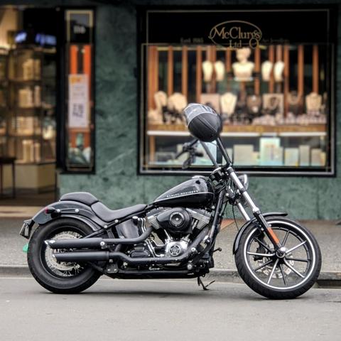 A slick black Harley-Davidson Softail motorcycle, parked on a city sidewalk against the backdrop of a jewellery shop storefront. The bike is detailed with blacked-out components, including the exhaust pipes and engine casing, and features a black helmet resting on the seat. The background storefront, with "McClurgs Ltd." clearly visible, has a distinctive green marble facade.