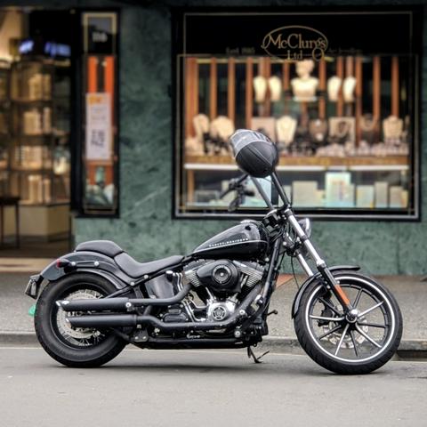 A slick black Harley-Davidson Softail motorcycle, parked on a city sidewalk against the backdrop of a jewellery shop storefront. The bike is detailed with blacked-out components, including the exhaust pipes and engine casing, and features a black helmet resting on the seat. The background storefront, with "McClurgs Ltd." clearly visible, has a distinctive green marble facade.
