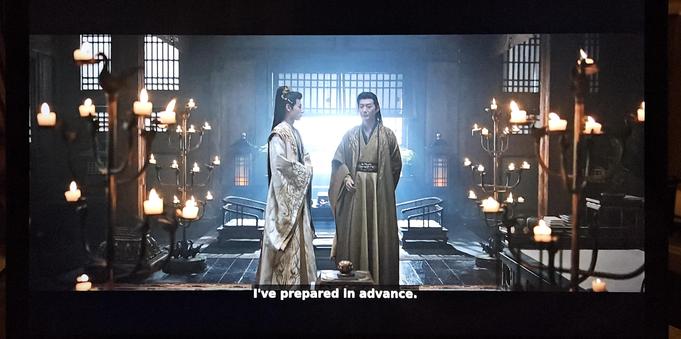 Two characters talking in a room with four candelabras. Each candelabre is almost two meters tall and have three levels of arms portruding from a pole in the middle. At the end of the arms and at the top of the poles sit stylised geese holding cups for the candles. The pole is fastened to a round stone foot, and on the foot four more geese are standing with their beaks against the pole.