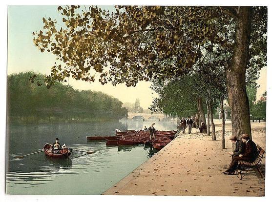 The image depicts a serene riverside scene with lush greenery and mature trees. A calm river flows through the center, reflecting soft light from an overcast sky above. Several boats are on the water; some appear to be moored while others have occupants rowing or sitting inside. On the right bank of the river is a paved promenade where several individuals engage in leisure activities such as walking and sitting on benches.

In particular, one area captures two men seated on a bench engaged in conversation with each other. Near them stands another man leaning against a railing, observing his surroundings. Further along the path, more people are seen strolling or standing at various points, enjoying their time by the water's edge. The overall ambiance suggests an early 20th-century setting, as indicated by historical attire of the individuals and vintage postcard style presentation.

The background features architectural elements such as bridges spanning across the river, connecting different parts of the town on opposite banks. This picturesque environment embodies a tranquil riverside atmosphere typical of leisurely towns during that period in England.