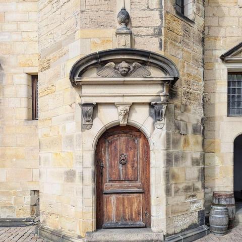 A dark wooden, arched castle door set in a stone wall, topped by a detailed stone carving of an angel and flanked by old wooden barrels.