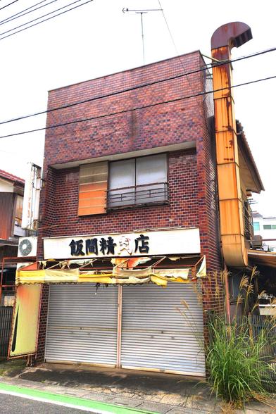 A boxy looking two-storey building that at least looks like it's built with bricks. At the bottom is a storefront whose shutters are down and the butchery name sign above it is in advanced decay. The plastic orange awning is in tatters. There is a smokestack running alongside the building on the right that's gotten rusty. The single window on the upper floor is covered by white tarp.