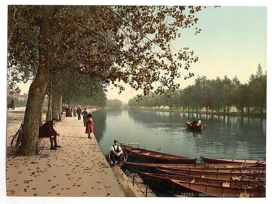The image depicts a serene riverside scene, possibly from an early 20th-century postcard or painting. In the foreground, on a tree-lined promenade with fallen leaves scattered around, several individuals are engaged in leisurely activities such as sitting and walking beside the water's edge. A person is seen seated by a bicycle near a bench under large trees, while another figure sits comfortably at a small table along the path.

In the middle ground, across calm river waters reflecting hues of greenery on their surface, several boats are moored or in use; one with passengers appears to be rowed and navigated through the waterway by an individual wearing traditional attire. The background showcases dense trees lining the far side, suggesting a park-like setting.

The overall color palette is subdued yet rich with earthy tones, capturing what seems like late autumn ambiance. A soft glow envelops the scene, hinting at early morning or late afternoon lighting conditions. This picturesque riverside tableau exudes tranquility and captures everyday life in an English urban environment from about a century ago.