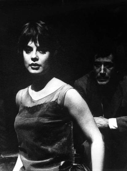 The photograph is a black and white image depicting two individuals. The person in the foreground appears to be sitting with their body turned slightly towards someone or something off-camera, as indicated by the positioning of one arm behind them for support on what seems like a chair. They have shoulder-length hair that falls just past the shoulders, styled straight and parted on the side.

Their expression is serious but not overtly emotional; they seem to be in deep thought or focused attention. The individual's clothing consists of a sleeveless top with visible straps over another garment, suggesting casual attire possibly from an earlier decade given the style. 

The lighting highlights only one side of their face and body, creating strong shadows on the other side which adds drama to the image.

In contrast, the second person is in the background, slightly out-of-focus due to depth of field technique typically used by photographers to draw attention primarily towards the subject in focus while still including context or another character. This individual appears older with a more rugged appearance; their face shows signs of aging such as wrinkles and deeper-set features.

They are wearing what looks like a dark jacket, suggesting a cooler environment or perhaps an outdoor setting where they might be involved in some activity that requires protection from the elements. The lighting on this person is less pronounced [...]