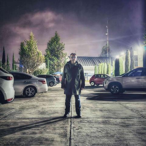 Un hombre vestido con una gabardina verde está de pie en un estacionamiento, entre varios autos. El cielo nocturno es púrpura brillante.