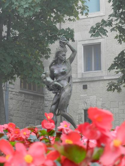 A photograph taken in San Marino shows the bronze “Monumento alle vittime del bombardamento, 26 giugno 1944.” The sculpture depicts a dramatic, dynamic female figure stepping forward with wind-swept hair raised in one hand, symbolizing strength and anguish. Her other arm holds a limp child against her torso, representing the civilian victims of the 1944 bombing. The statue stands before a stone building with rectangular windows and is partially framed by lush green trees. In the foreground, out of focus, a dense cluster of bright pink and red flowers adds vivid color and soft contrast to the somber monument. The misty, muted background enhances the emotional weight of the scene, emphasizing remembrance and loss.