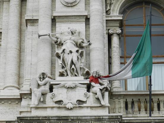 A detailed photograph of the façade of the Corte di Cassazione in Rome, taken on October 9th, 2012. The image focuses on an ornate section of the pale beige travertine building, where three sculpted figures stand above a row of classical columns. In the center, a majestic female figure in flowing robes raises a long trumpet to her lips, symbolizing justice or proclamation. Below her, two cherub-like figures sit on either side of an elaborate stone ledge. The cherub on the left leans against a shield, while the one on the right reads from an open book draped with the Italian flag, whose red, white, and green fabric billows outward toward the right side of the frame. Behind the sculptures, a tall arched window with wooden framing contrasts with the carved stone. The architectural details—scrollwork, decorative reliefs, and vertical columns—convey the grandeur and solemnity of Italy’s highest court of appeal.