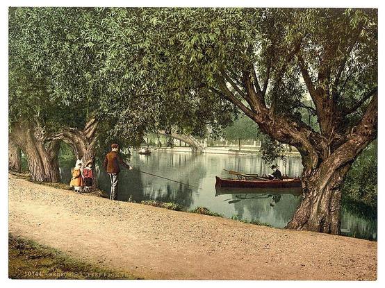 The image captures a serene riverside scene. A man in casual attire is standing on the left side, engaged in fishing with a rod that extends into the river's surface. He appears focused and relaxed as he casts his line. To his right, two children are seated on what seems to be a boat-like structure or perhaps an oversized chair by the water’s edge. One child has blond hair adorned with orange flowers, likely roses, suggesting springtime activities, while the other sports red clothing paired with white leggings.

On the opposite bank of the river, another individual is seen in more formal attire, possibly early 20th-century fashion, seated on a wooden boat and navigating its waters. This person seems to be enjoying leisurely rowing or drifting down the calm expanse of water that reflects muted hues from overcast skies above.

The backdrop features lush foliage consisting primarily of broadleaf trees with rich green leaves suggesting full bloom during late spring to early summer months, which enhances the tranquility of this idyllic setting. The riverbank is lined with a path made visible in shades of brown and tan dirt, inviting for strolls or leisurely walks.

The overall ambiance conveys an era where such pastimes were commonplace—highlighting simplicity, nature’s beauty, and recreational activities that typified daily life during the late 19th century. The image is marked by a vintage aesthetic with its faded colors, i [...]