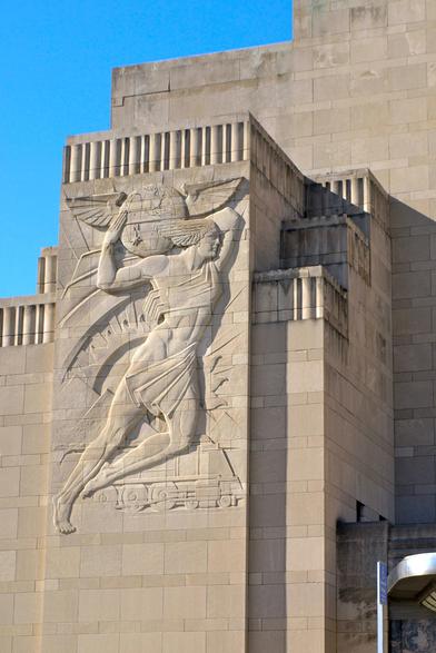 A large, detailed bas-relief sculpture is set into the side of a massive, tan-colored, stepped architectural structure, likely a building facade. The sculpture depicts a figure in motion, appearing to be running or leaping forward with arms raised. The figure is highly muscular, wearing a simple garment and what appears to be a winged helmet. Below the figure is a cluster of smaller sculpted elements,representing a railroad locomotive. The architectural structure behind the relief is characterized by repeating rectangular blocks and stepped levels, creating a strong sense of geometric form and depth.