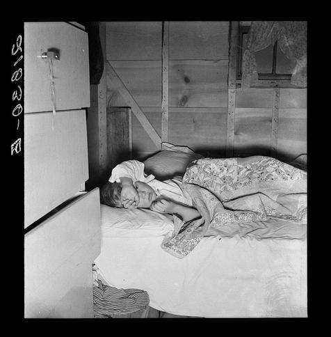 This black and white photograph depicts a young child lying on an unmade bed within what appears to be the interior of a trailer. The setting suggests modest living conditions with wooden walls, a window draped partially by curtains, and minimal furnishings aside from the mattress where the child rests. A refrigerator is visible in the foreground, indicating limited space for food storage or preparation.

The expressionless face of the sleeping child conveys vulnerability and potential discomfort. The overall atmosphere of the image evokes a sense of hardship or struggle associated with rural life during what could be interpreted as a historical period of migration and economic challenge, particularly given the mention of "Farm Security Administration" (FSA), which was part of efforts to support agriculture in America.

The photograph is labeled with handwritten annotations indicating it's from an FSA mobile camp. The specific location mentioned is Merrill, Klamath County, Oregon, suggesting a connection to rural communities and their living conditions during the time period when such images were commonly produced for documentation purposes by government programs like FSA that focused on farm workers' lives.

The visual elements of the photograph—the child's peaceful repose against a backdrop of sparse, utilitarian surroundings—could be interpreted as reflecting themes related to migration, survival, and resilience withi [...]