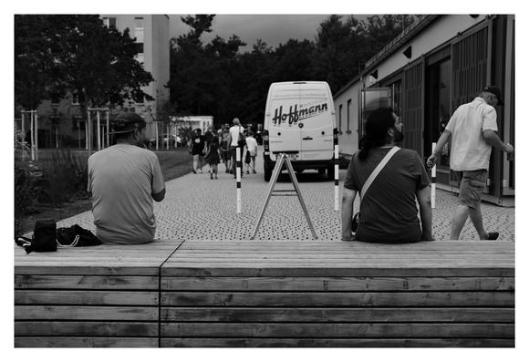 Schwarz-weiß-Szene in einem urbanen Außenbereich: Zwei Personen sitzen auf einer Bank, eine dritte geht vorbei; im Hintergrund ein Van mit der Aufschrift „Hoffmann“ auf Kopfsteinpflaster.
