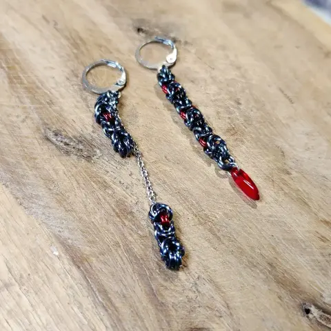 Boucles d'oreilles en maille, asymétriques, dans des couleurs gris et rouge. L'une des boucles est longue et se termine par une perle en forme de goutte de couleur rouge, et l'autre est en deux parties : une courte, et une plus longue attachées à une petite longueur de chaînette.