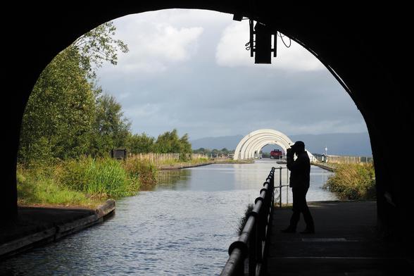 Blick aus einem Tunnel auf einen Kanal, der auf eine Schiffshebebühne zuführt. Im Tunnel, der einen dunklen Rahmen für das Foto bildet, steht ein Mensch mit einer Kamera in der erhobenen Hand. Der Himmel ist wolkenverhangen, blaugrau,  links des Wassers wachsen Bäume und Büsche.  Im Hintergrund die weißen halbkreisförmigen Bögen des Falkirk Wheel, durch die ein Schiff fährt.

View from a tunnel onto a canal leading to a ship lift. In the tunnel, which forms a dark frame for the photo, stands a person with a camera raised in their hand. The sky is overcast, blue-grey, with trees and bushes growing to the left of the water. In the background are the white semicircular arches of the Falkirk Wheel, through which a ship is passing.