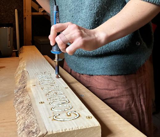 Piece of wood with letters "goodbye corner" burned in and filled with white goo. Person behind is using soldering gun to make some more marks into the wood.