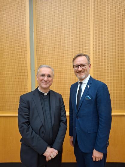 Vor einer hölzernen Wand stehen zwei Personen. Links steht ein Mann mit grauen Haaren und dunklem Anzug. Der Mann trägt einen sog. Priesterkragen. Rechts steht ein etwas größerer Mann in blauem Anzug und Krawatte. Beide Männer lächeln ins Bild. Bei dem Mann links handelt es sich um Stefan Heße, Erzbischof von Hamburg. Bei dem dem Mann rechts handelt es sich um Sebastian Holzbrecher, Professor am Institut für Katholische Theologie an der Universität Hamburg.