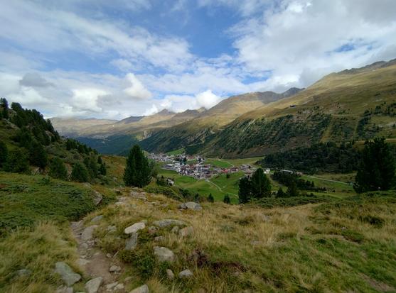A hiking trail amidst grass and trees. Down below is the small village called Obergurgl. The valley opens up to the viewer.