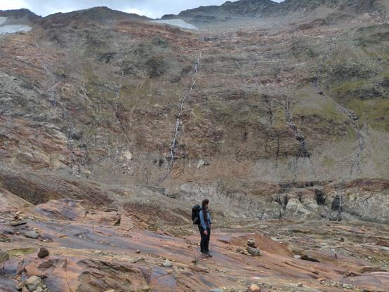 A woman with a blue jacket and backpack stands on a large rock and looks to thge side. A high-rising rock wall can be seen behind her. Small water streams trickle down the wall.