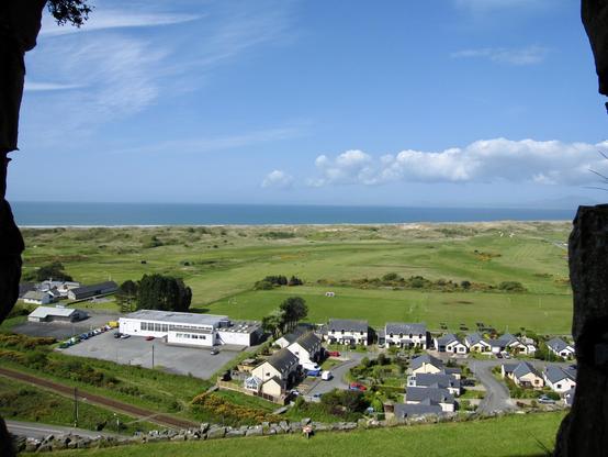 A small coastal settlement is framed by rocky edges, with a cluster of houses and a few larger buildings surrounded by green fields. Beyond the houses lie open grassy areas with scattered football pitches and a golf course stretching towards sandy dunes. The sea meets the horizon under a blue sky with scattered clouds, and distant hills are faintly visible across the water. A railway line runs near the lower edge, and the scene transitions smoothly from the compact village to expansive natural landscapes.