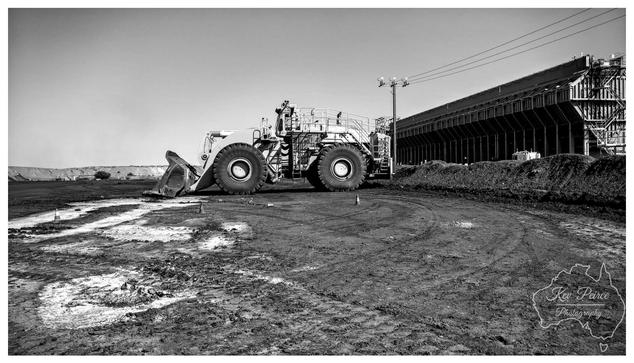 A wide view, black and white photograph signed by Kev Peirce. A massive front end wheel loader is positioned centrally on a heavily churned, muddy, and dark earth surface.  Its large bucket rests on the ground to the left. To the right, a long, dark industrial structure, possibly a conveyor system or processing plant, dominates the background.  A utility pole stands near the structure. The composition emphasises the heavy, industrial nature of the site and the contrast between the machine and the facility.