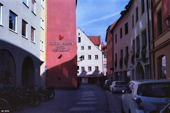 Farbfoto, das eine Straße in Regensburg zeigt. In Bildmitte die dunkelrosa Wand des Hotels Roter Hahn. Starker Schattenwurf im linken Teil des Bildes. Leica R4 auf Kodak Ektar.