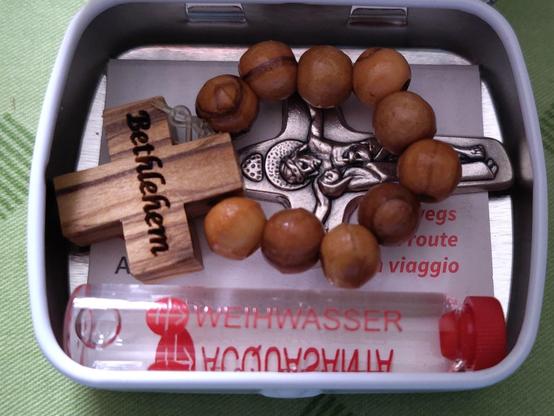 Hosentaschenkleine Alubox des Hilfswerks "Kirche in Not", mit einem olivenhölzernen Fingerrosenkranz aus Bethlehem, einem backaromakleinen Fläschchen Weihwasser und einem Einheitskreuz-Anhängerchen aus Metall. Darunter liegt ein Zettel mit kath. Grundgebeten, in den drei Schweizer Landessprachen (DE/FR/IT). Die Box ist in weisser Grundfarbe gestaltet, mit signalroten Schriftakzenten.
