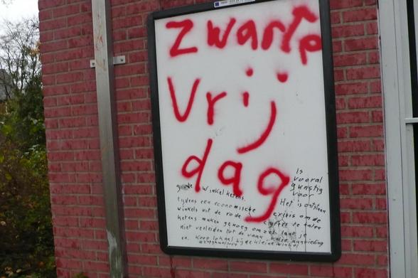 bakstenen muur met metalen lat er op en rechts een deel van een deur kozijn.
Reclame frame aangepast met witte poster met rode spray letters en zwarte tekst als in caption. rechts van de muur wat groen begroeing