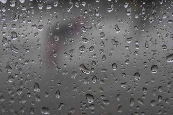 A close-up view of a rain-speckled surface, likely a window, covered in numerous water droplets of varying sizes. The droplets create a textured, abstract pattern, reflecting muted light and subtle shadows. The background is blurred, drawing focus to the intricate details of the raindrops clinging to the glass, evoking a moody and contemplative atmosphere.