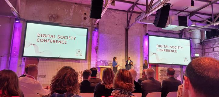 A stage with three speakers in a factory-like setup with two large screens showing the words "Digital Society Conference"