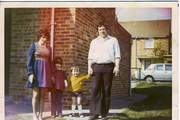 A scan of a 1970s colour photo. The colours are a little washed out and there are indications of light leaks in the camera body affecting the final photo, particularly along the top edge and top right. The image is taken outside a brick house on a sunny day, at its rear on the corner. As the house sits at the end of a small road it has grass to the right forming an extended garden barrier before the pavement to the right out of sight and in the background behind the side of the house. There are other terraced houses across the road to the rear and the back of a silver car can be seen. Standing to pose for the photo are two adults either side of two children, everyone holding hands. The woman on the left has dark hair and a dress that comes to mid-thighs, in dark blue material for the main and down the full sleeves but patterned with thin yellow and white bands across the torso area. The man on the right also has dark hair and wears dark, slightly flared trousers with a white shirt with sleeves rolled up a little. Both adults have cigarettes in their hands. Between them is a girl and a boy, both young but the girl is slightly taller and older. She has blue trousers and a red and purple top. The boy has a yellow jumper, blue shorts, long white socks, and blue sandals. The children are smiling while the parents look a little resigned.