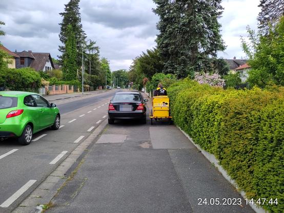 Eine briefzustellende Person quetscht sich mit dem Postfahrrad an einem auf dem Gehweg parkenden PKW vorbei.