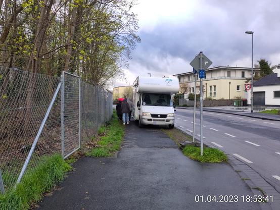 Eine Grupe von Fußgänger*innen läuft im "Gänsemarsch" an einem auf dem Gehweg abgestellten Wohnmobil vorbei.