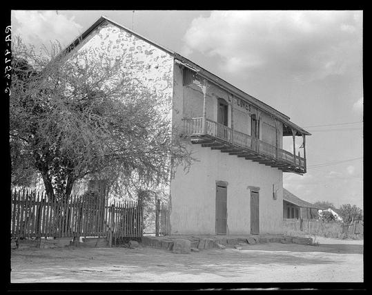 The image depicts a two-story house with signs of significant wear and decay. The building is painted white but appears to be peeling in several areas, revealing the underlying surface or brickwork. It features wooden railings on both floors around an open balcony on the second floor, which has no roof covering it. There's visible damage like cracks near one corner where a door used to stand and some of these structural issues are consistent with older architecture that may have been neglected over time.

In front of the house is a rustic-looking wooden fence partially enclosing what seems to be an outdoor area or yard. The ground in this vicinity appears dry and uneven, possibly due to lack of maintenance. There's also a cluster of trees on the left side providing some contrast with their dark silhouettes against the sky. Above the building, power lines stretch into the distance under a cloudy sky.

The monochromatic tone suggests that it could be an older photograph or taken in black and white for stylistic reasons to emphasize texture over color. The text at the bottom of the image indicates "Dorothea Lange," which is associated with her work during the Great Depression documenting social issues, including poverty among Mexican-American families on the American-Mexican border.

This specific house appears within a broader context likely related to cultural or historical studies concerning rural Texan communities and t [...]