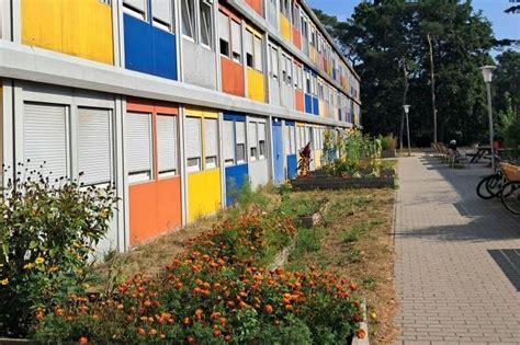 Links ein dreistöckiger, bunter Container-Bau einer Geflüchtetenunterkunft. Davor ein Grünstreifen und rechts ein gepflasterter Fußweg