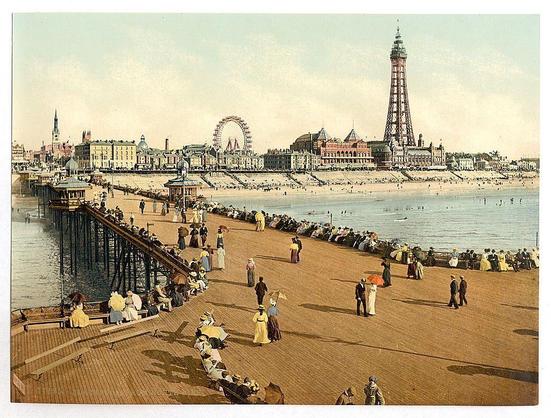 The image depicts a vibrant and bustling seaside scene, likely from late 19th to early 20th century England. It showcases the North Pier in Blackpool with its iconic architecture prominently featured on the right side of the frame. A tall tower-like structure stands out against the sky, symbolizing an era where such architectural feats were a source of pride and fascination.

The beach area is teeming with people engaging in various activities typical for that time period: walking along the promenade, sitting under parasols or gazebos, conversing in groups on benches. The attire suggests a warm season, as individuals are dressed in light, summer-appropriate clothing, including dresses for women and suits for men.

In the background across the water lies an array of buildings that appear to be part of Blackpool's entertainment district, with one particularly large structure featuring what looks like Ferris wheels or observation towers. The cityscape is a mix of Victorian-era architecture alongside more modern designs, hinting at the area’s evolution over time and its status as a major resort destination.

The overall color palette includes soft earth tones for the buildings and boardwalk, contrasted by bright yellows in women's dresses, reds from parasols or clothing, and the natural hues of the beachgoers' outfits. The photograph has been preserved with visible wear, including faded colors and creases, indicative of its  [...]