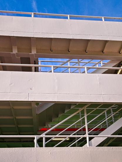 The stairs of a ship seen from the side. Two floors are visible, everything is painted white, including the railings. In the background, part of the ship is painted green. Blue sky can be seen through the top floor and the top deck. Heavens, this is difficult to describe. It's mainly metal and stairs. And a cloudless sky.