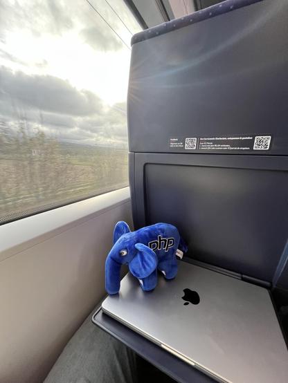 PoV from train seat. Left half is view out the window with fields and a cloudy sky. Right side it the seat in front with a MacBook on the tray and a blue PHP elephant on top of it.