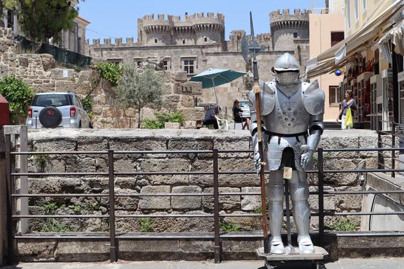 Eine metallene Ritterstatue steht in der Nähe einer Steinmauer in einem historischen Viertel, im Hintergrund ist ein burgähnliches Gebäude zu sehen. In der Nähe ist ein weißes Auto geparkt, und Menschen sitzen unter einem Sonnenschirm im Freien.