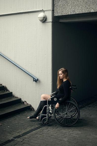 Eine junge Frau sitzt in einem Rollstuhl vor einem dunklen Durchgang. Sie trägt schwarze Kleidung und hat an beiden Beinen Gipsverbände, eins reicht bis zum Knie, das andere bis ganz nach oben. Die Frau ist seitlich zur Kamera gedreht, ihre Haare sind schulterlang und hell. Im Hintergrund ist eine Treppe mit einem Handlauf an der Wand zu sehen, die nach oben führt. Die Wand ist hell und hat eine runde Außenleuchte angebracht.