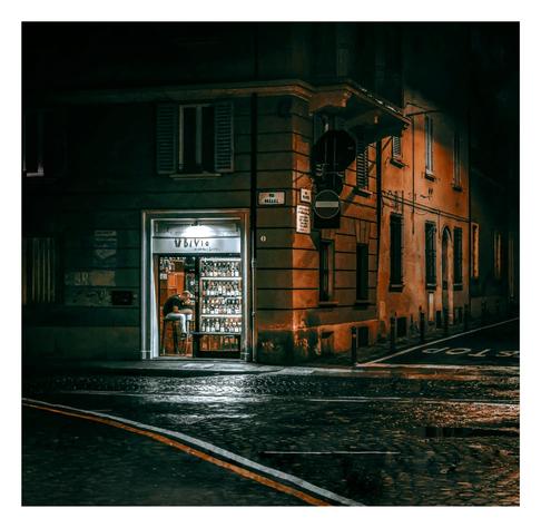 A photo of a dimly lit, narrow street corner at night. A small, warmly lit wine shop with a "Bivio" (the crossroads) sign is centered, its display window filled with various winebottles. The building is aged, with weathered brown/orange and grey walls, shutters, and a streetlight casting orangey light. The wet cobblestone street reflects the light. The background is shadowy, with faintly visible neighboring buildings.