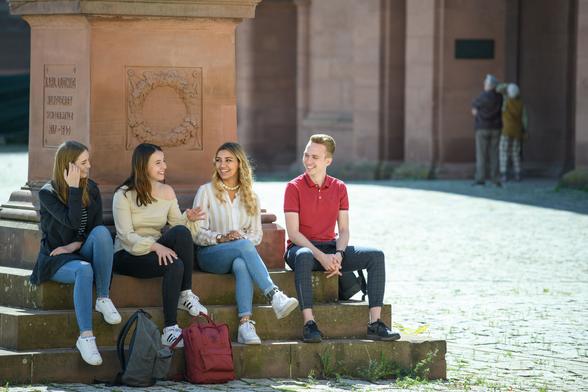 Vier Studierende sitzen auf den Stufen einer Statue auf dem Ehrenhof. Sie lachen.