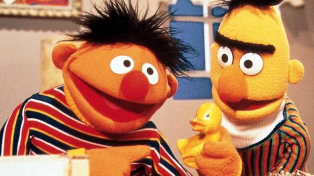 La imagen presenta dos coloridos personajes de marionetas con peinados distintos y ropa a rayas. Un personaje (EPI) sostiene un pequeño pato de goma amarillo, mientras ambos sonríen e interactúan entre sí. El fondo incluye una ventana y obras de arte enmarcadas.