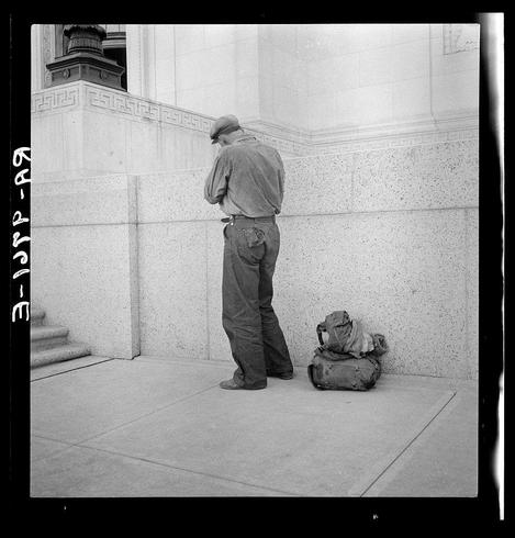 This black and white photograph depicts an individual standing in a public space with their luggage. The person appears to be dressed casually, wearing pants and a jacket suitable for mild weather conditions, indicative of a worker who might travel frequently or live on the streets without permanent shelter. They are leaning against what seems like a large stone structure, possibly part of a building's exterior wall.

Two pieces of luggage accompany them: one is placed upright with its handles visible, suggesting it may be in use for immediate transportation needs, while another lies nearby on the ground, partially obscured from view but clearly intended as travel baggage. The setting appears to be an urban environment due to architectural elements like stone and concrete commonly found around city buildings.

There's a sense of solitude conveyed by their posture—leaning against the wall with bags close by yet no other individuals within sight. This can imply several scenarios such as waiting for transport, taking a break from travel or laborious work that might demand frequent relocation between sites.

No context regarding race is apparent in this image; it portrays an individual without specifying racial identity but does evoke themes of poverty and displacement through the juxtaposition of belongings with homelessness against urban architecture.