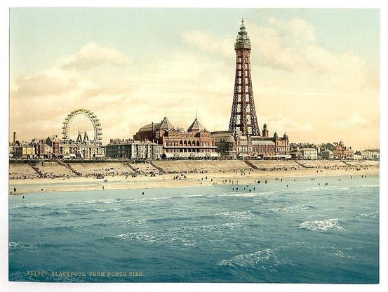This image depicts a vibrant and bustling beach scene from North Pier in Blackpool, UK. The composition showcases the iconic observation tower known as "The Tower," prominently rising against an overcast sky with hints of orange hue suggesting either sunrise or sunset lighting conditions. In the foreground, we observe shimmering blue waters where people appear to be enjoying various activities such as swimming and wading.

In contrast to this lively atmosphere on land, a large Ferris wheel is visible in the background, adding another layer of amusement park-like excitement to the setting. The beach itself seems to have multiple levels or terraces with steps leading down towards the shore, indicating designated areas for visitors' use. A dense collection of buildings lines the coastline behind the shoreline, displaying architectural styles that suggest a historical context from approximately late 19th century England.

The overall ambiance is one of leisure and pleasure typical of seaside resorts during this era in Britain. The image captures not only physical elements but also an essence of time—a snapshot of public life at North Pier around Blackpool between the early to mid-20th century, where people gathered for entertainment and relaxation by the sea.