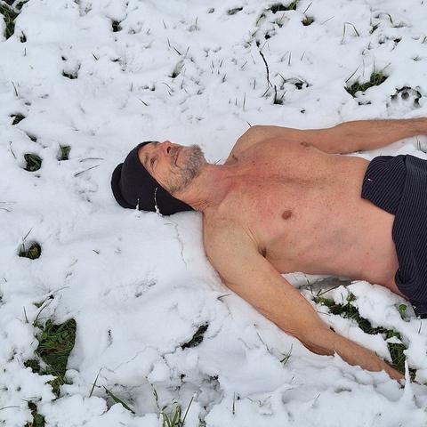 Der Oberkörper eines Mannes mit schwarzer Mütze. Er liegt unbekleidet auf dem Rücken im Schnee. Auf seiner Hüfte liegt ein dunkler Schal. Durch die dünne Schneedecke sind stellenweise grüne Gräser zu sehen.