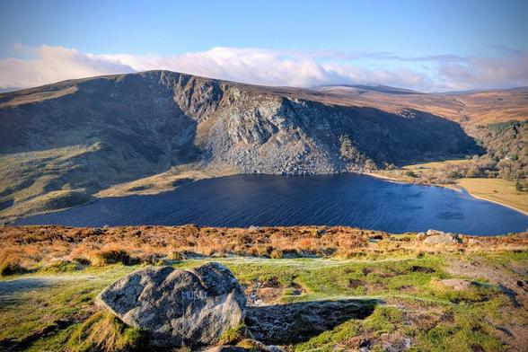 A breathtaking wide shot of Lough Tay in the Wicklow Mountains, Ireland. A dark, deep-blue lake is nestled at the base of a steep, rugged, rocky mountain face. The foreground features a grassy slope with patches of frosty, golden-brown and green vegetation, and a large, moss-covered boulder with carved initials. The sky is clear blue with high, wispy clouds catching the sunlight, and the distant hills are covered in heath and woodland. The mood is tranquil and expansive.