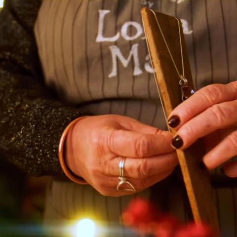 Close-up of some hands putting a piece of jewellery on a wooden stand. The LollaMac logo can be seen on an apron, and there is a blurry fairy light and some berries in the foreground.